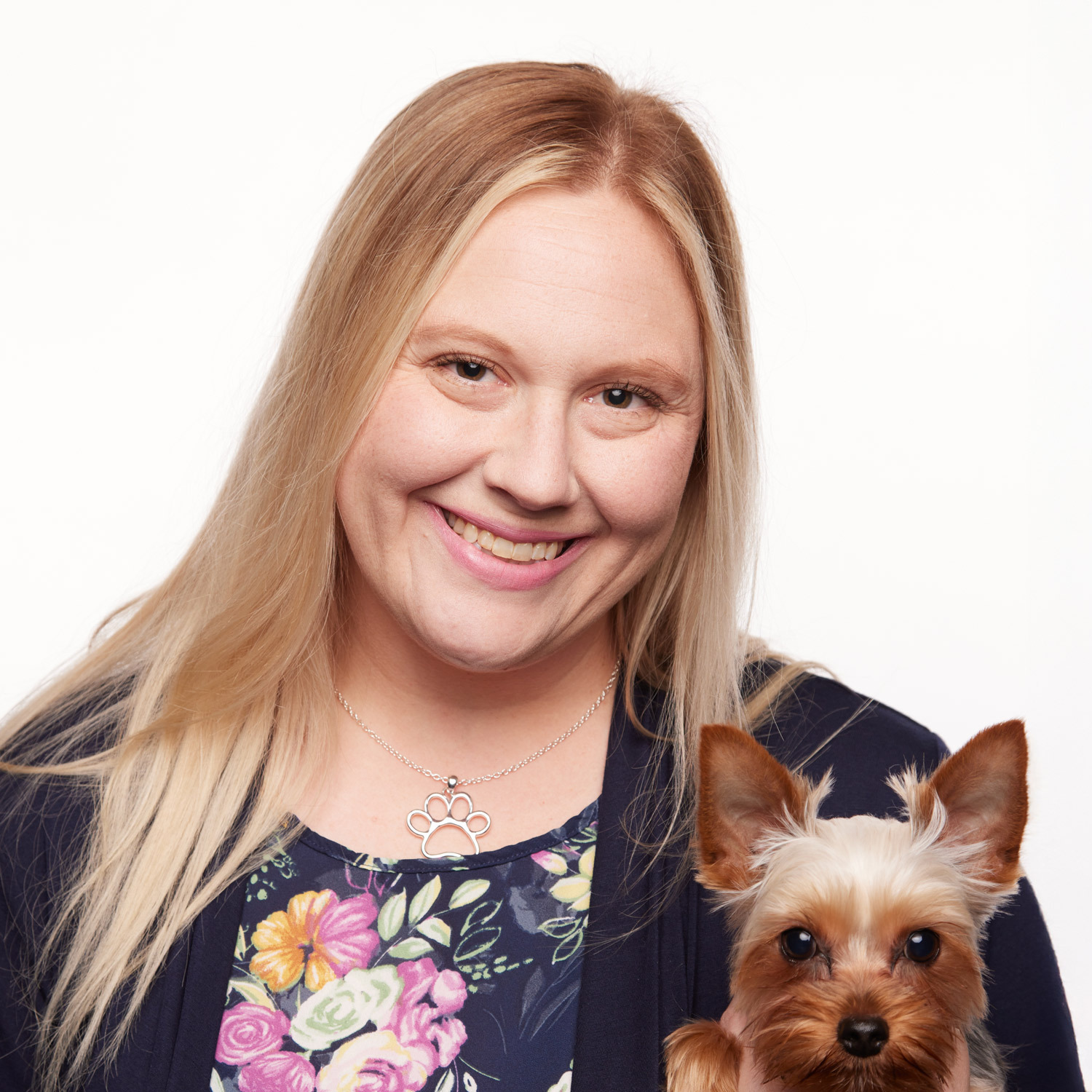 This image depicts a blonde woman with a floral shirt holding a fluffy dog smiling at the camera.