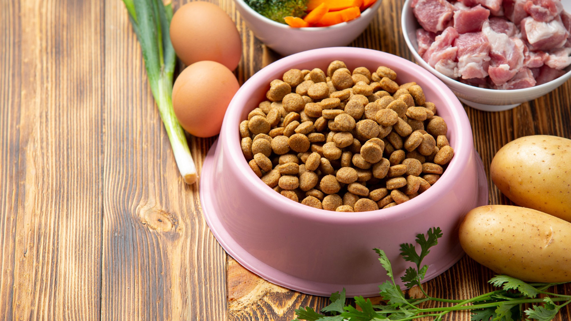 This image depicts pet kibble in a pink bowl next to eggs, potatos, meat cubes, an herb and leeks.
