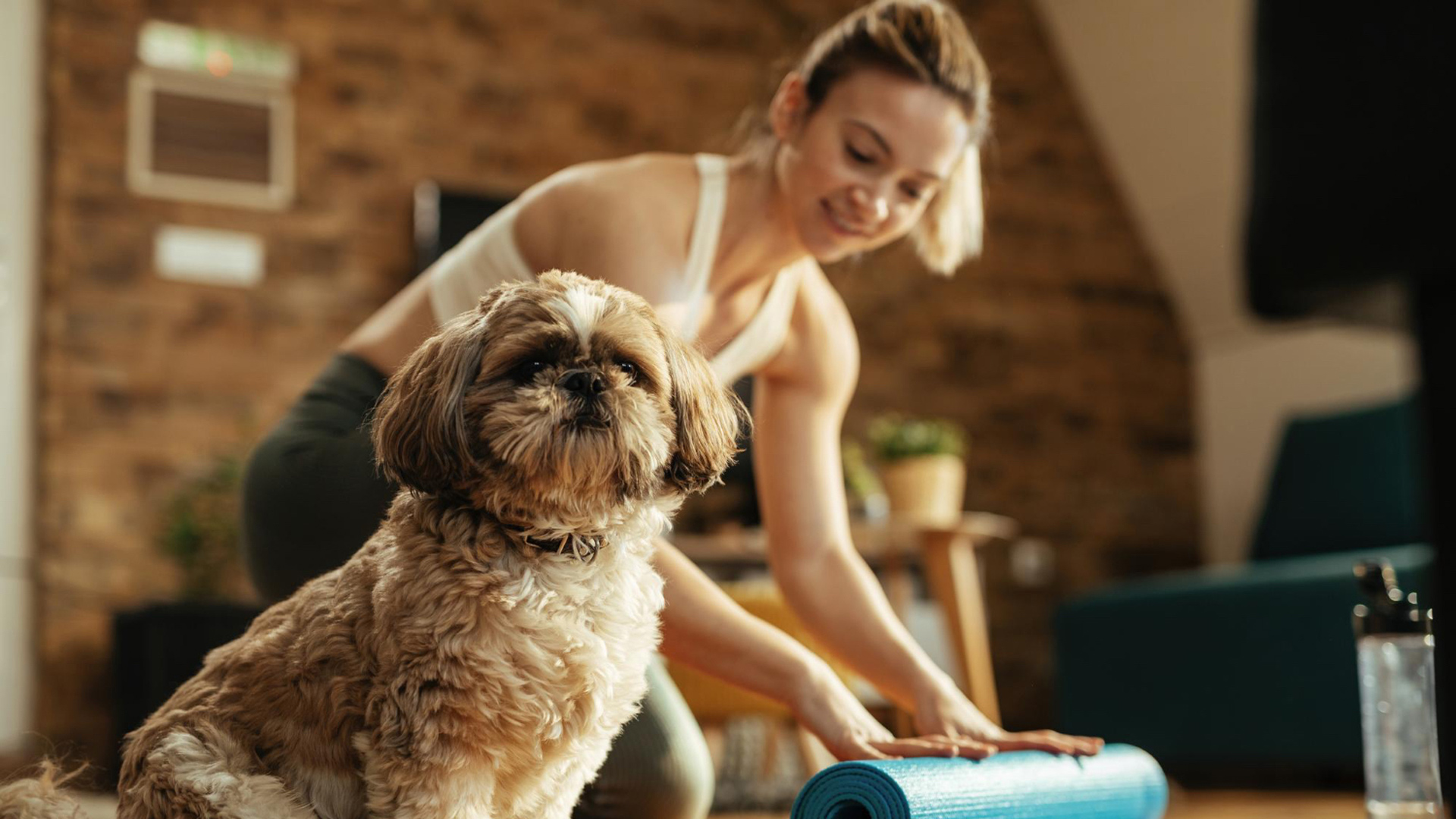 This image depicts a woman rolling up a blue yoga mat with a fluffy dog on the ground.