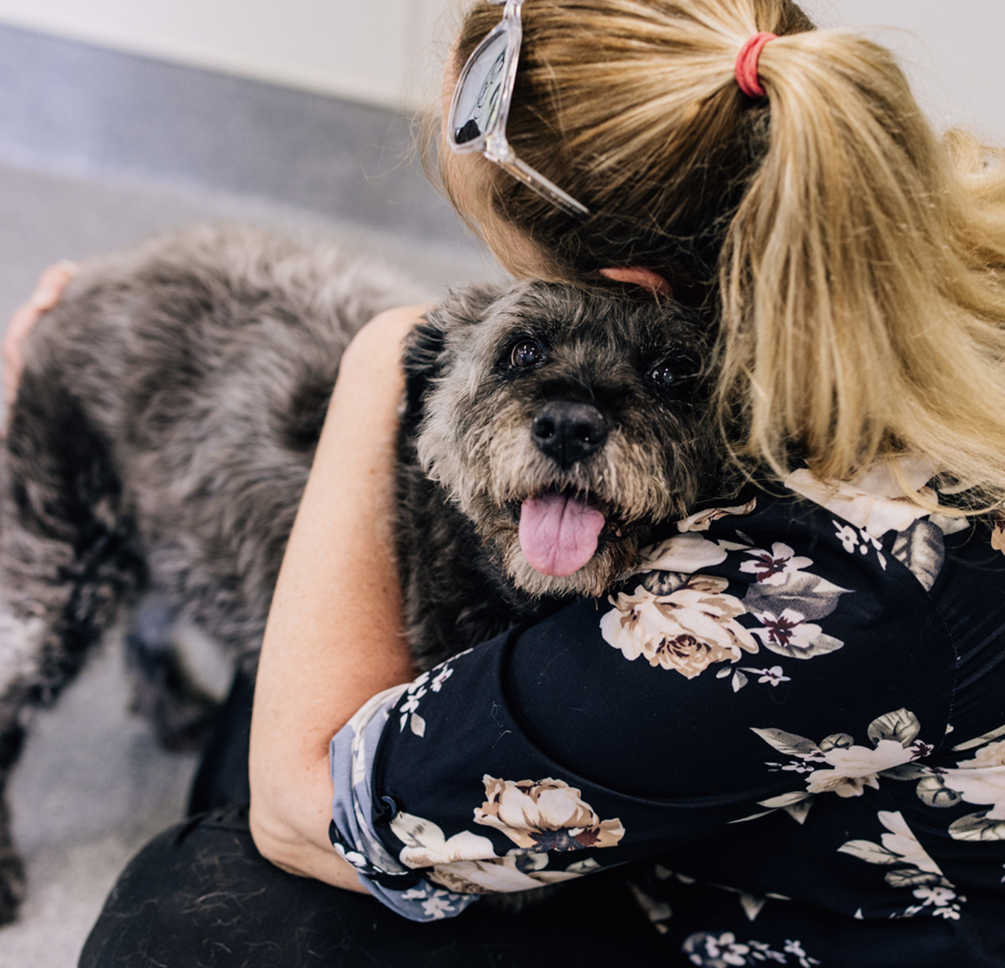 This image depicts a woman with a floral shirt hugging a black dog.